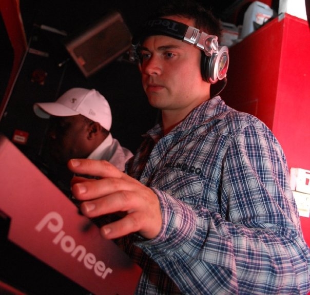 Russell Ruckman djing behind a red wall, with a friend wearing his hat